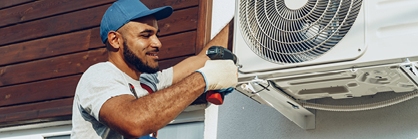 A man wearing blue coveralls and a blue hat stands on a ladder to install a fan.